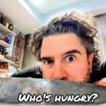 Close-up of a man raising one eyebrow behind a chef’s knife with the words “Who’s Hungry” printed on the blade